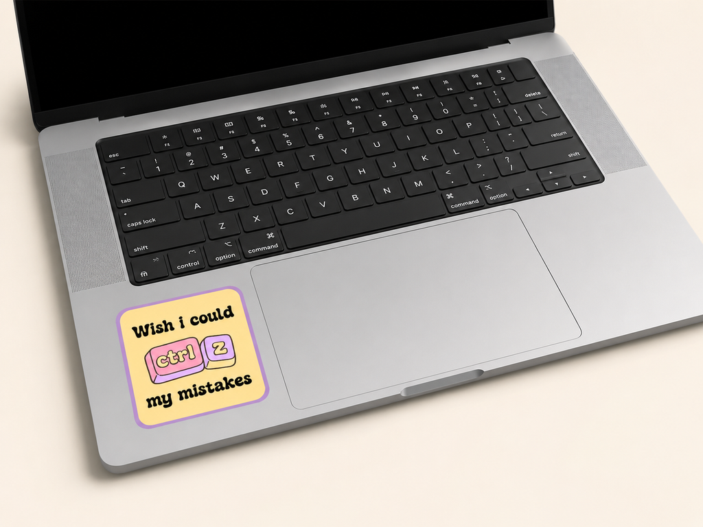 A silver laptop with a black keyboard on a light beige background, featuring a square sticker on the palm rest that reads 'Wish i could ctrl + z my mistakes' with pink and yellow keyboard keys.