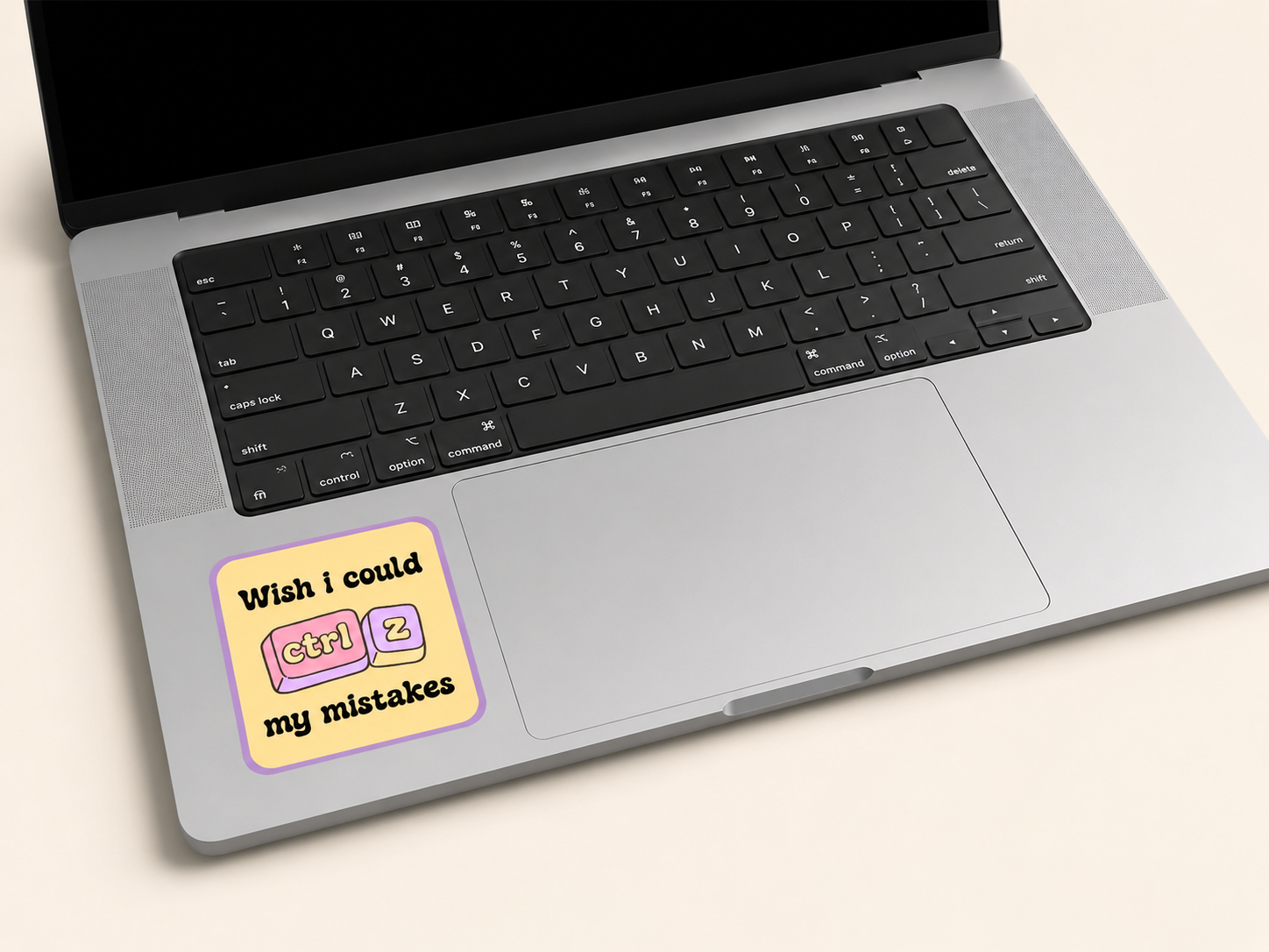 A silver laptop with a black keyboard on a light beige background, featuring a square sticker on the palm rest that reads 'Wish i could ctrl + z my mistakes' with pink and yellow keyboard keys.
