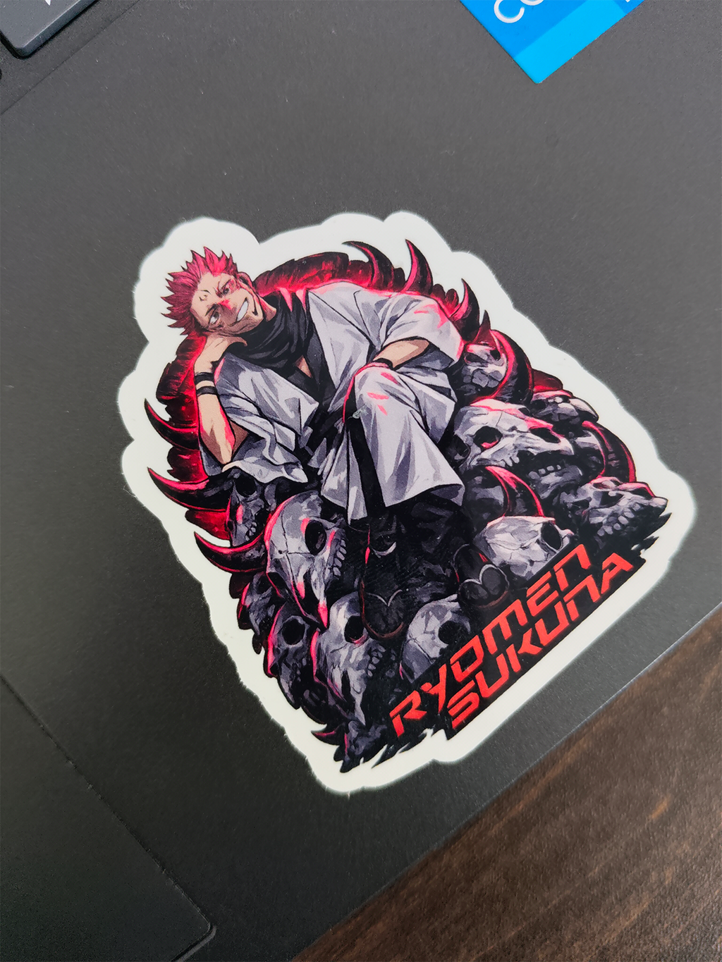 A die-cut vinyl sticker featuring an anime-style character with spiky red hair, a white gi, and dark gray skulls with red accents, placed on a dark laptop lid. The text 'RYOMEN SUKUNA' is printed in red at the bottom right.