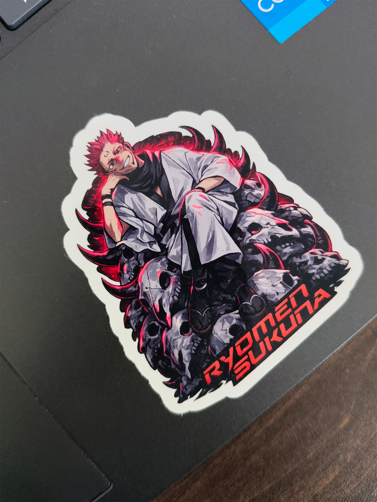 A die-cut vinyl sticker featuring an anime-style character with spiky red hair, a white gi, and dark gray skulls with red accents, placed on a dark laptop lid. The text 'RYOMEN SUKUNA' is printed in red at the bottom right.