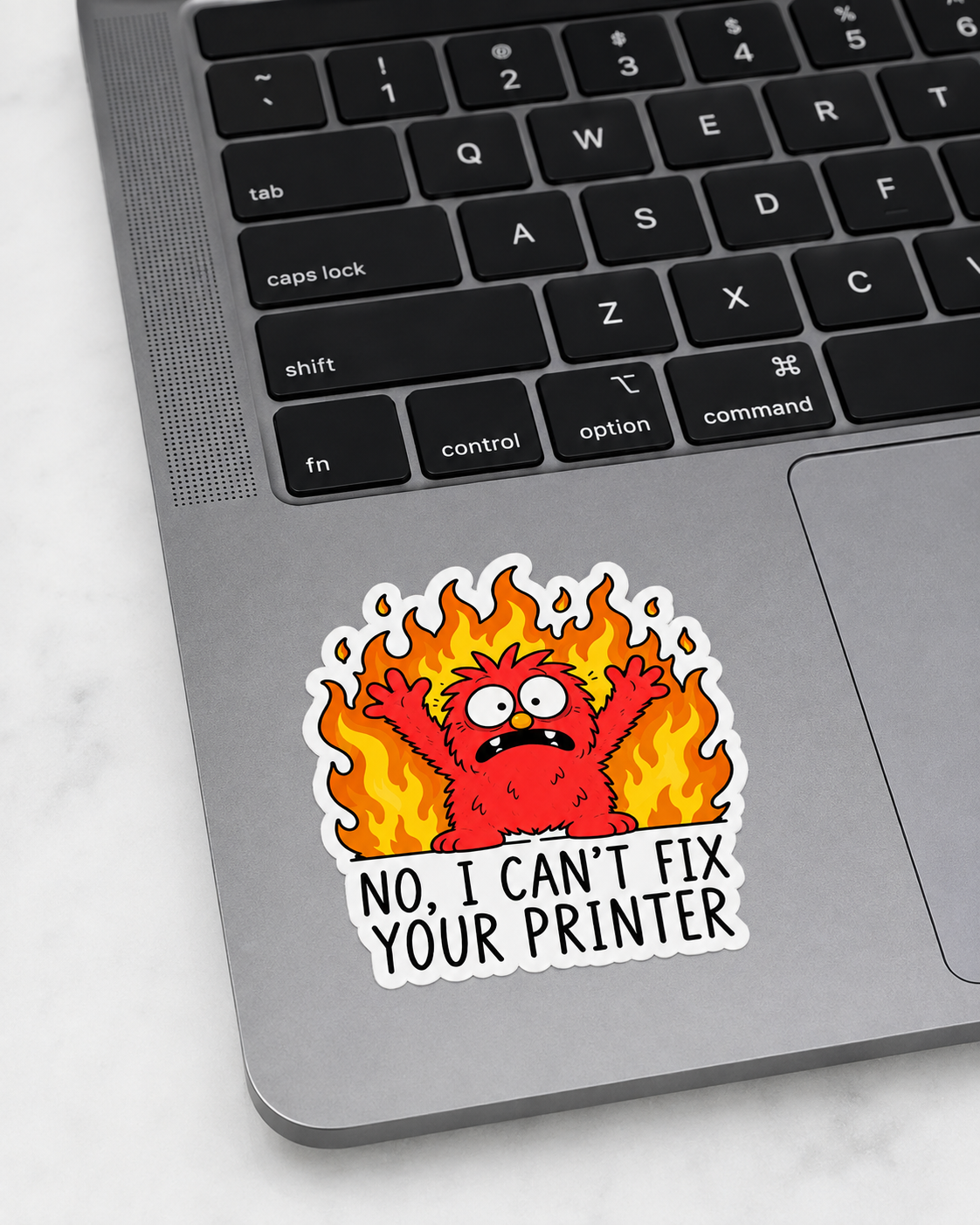 A die-cut vinyl sticker applied to the bottom corner of a silver laptop keyboard. The sticker features a cartoon red monster surrounded by orange and yellow flames, with the text "NO, I CAN'T FIX YOUR PRINTER" in black capital letters on a white background.
